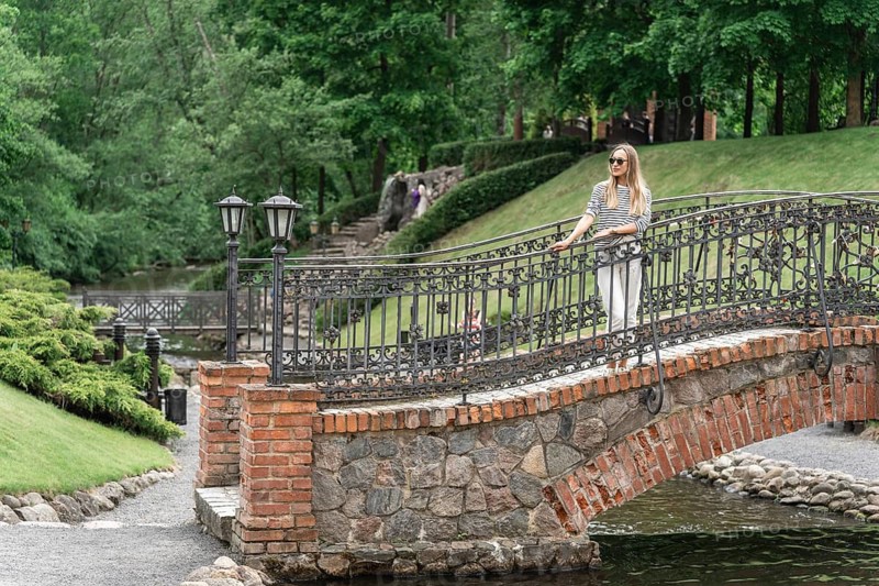 A woman stands on an old bridge in the park in the summer, Woman standing on a stone bridge with iron railing in a lush green park, creating a serene summer atmosphere, Belmontas Park, Vilnius, Lithuania
