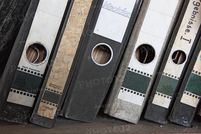Office scene around 1982, lost place, abandoned location, classic file folders on a wooden shelf, some of the folders are labelled in detail by hand and provide information about their contents, mixed up, dirty
