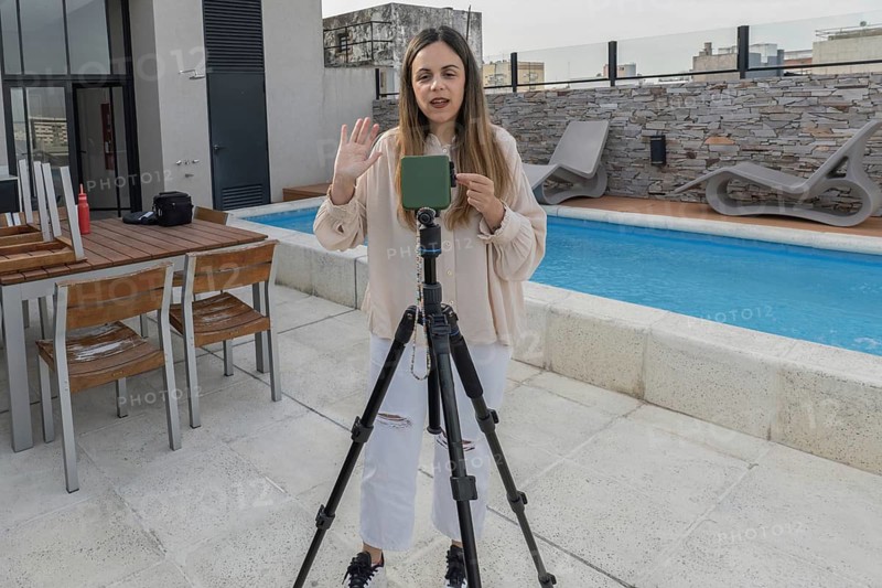 Woman standing behind tripod waving and talking to camera on rooftop terrace, wooden dining table and pool loungers visible around swimming pool
