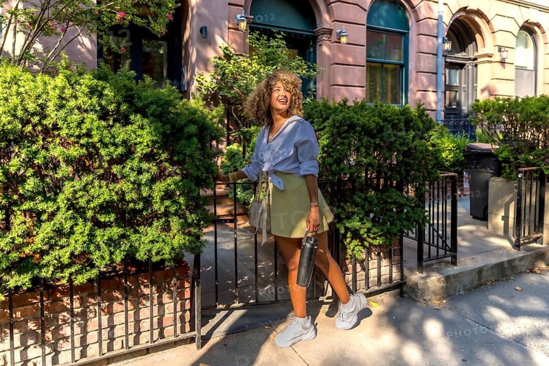 Woman with curly hair smiling and carrying a reusable bottle, walking out of a brownstone's gated green space on a sunny day, experiencing a healthy and active urban lifestyle in manhattan