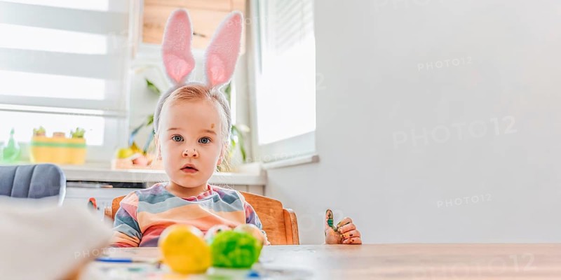 Banner with child wearing bunny ears sits at a table decorated for Easter. Colorful eggs are placed on the table. The room has bright light and festive items. Copy space for text