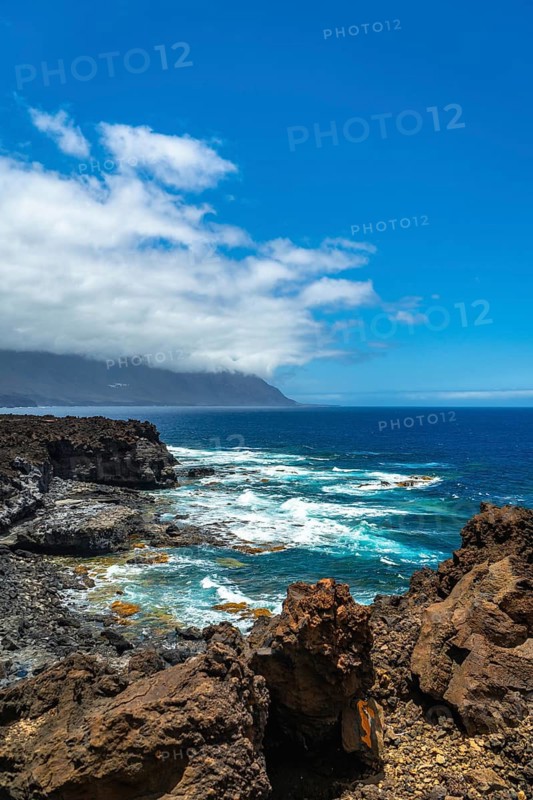 El hierro island's rugged volcanic coastline featuring dark rocks along the shore, clear blue ocean water with white crashing waves, under a partly cloudy sky with distant mountains