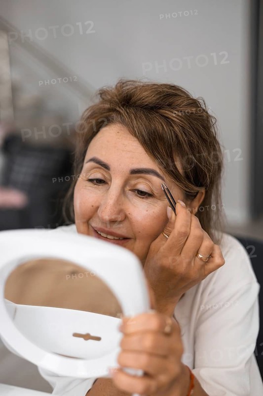 A woman smiles as she carefully shapes her eyebrows using a small brush while looking into a handheld mirror. The indoor environment is well-lit and inviting