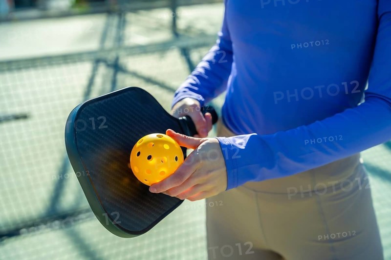 Person is holding a black pickleball paddle and a bright yellow wiffle ball, preparing to play on an outdoor court, symbolizing active recreation, healthy lifestyle, and skill in sports