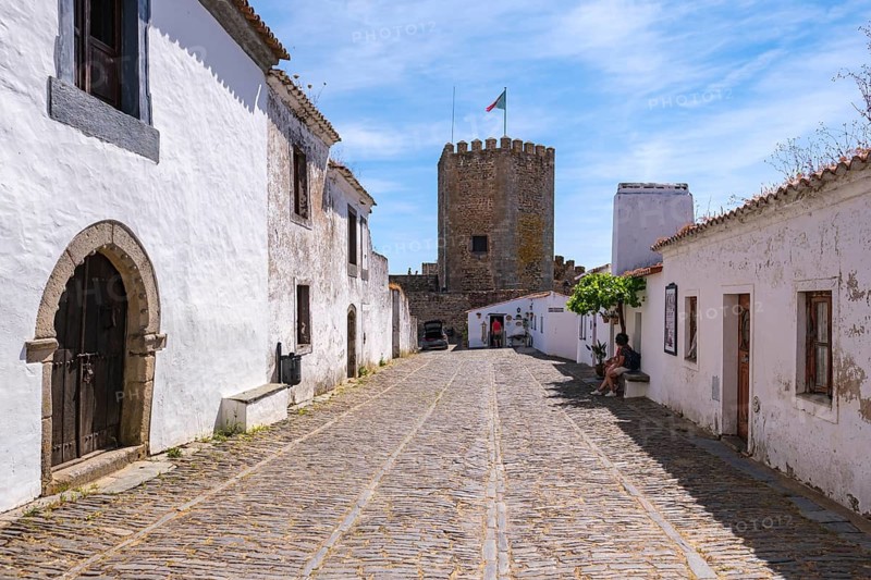 Paved street lined with historic white buildings and view of a castle tower, Monsaraz, Évora, Portugal
