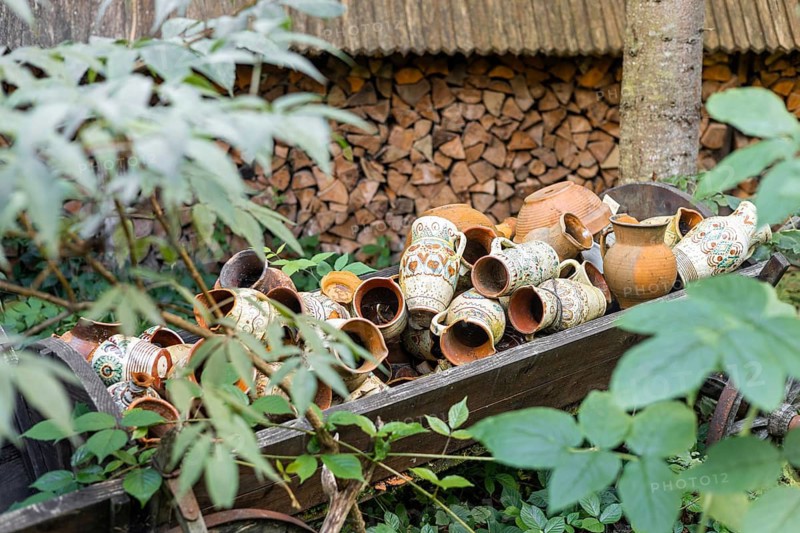 A collection of traditional clay pots in various sizes and designs, resting on a wooden cart surrounded by green foliage and stacked firewood in the background