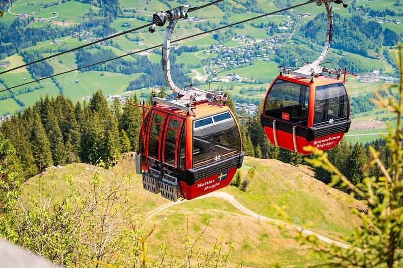 Two red cable car cabins float above a wooded hilly landscape with a wide view, Hahnenkamm, Kitzbühel, Austria