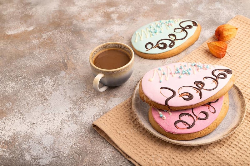 Glazed pink and blue decorated cookies, cup of coffee, on brown concrete background with linen beige textile, copy space, side view