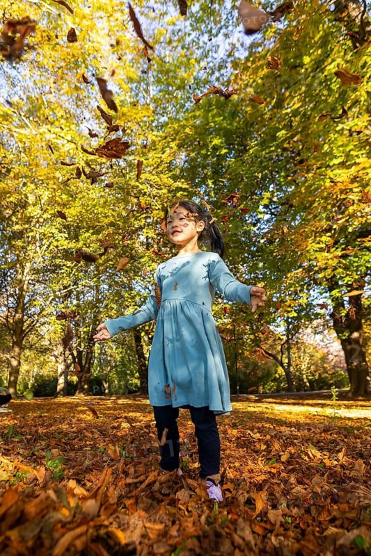 A joyful Asian child in a blue dress happily plays amid falling autumn leaves in a vibrant park setting, capturing the essence of carefree childhood and seasonal beauty