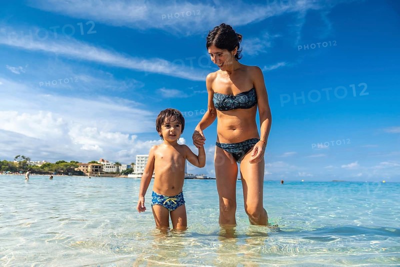 A woman and a child are walking in the ocean. The woman is wearing a bikini and the child is wearing a swimsuit. Scene is happy and carefree, as the mother