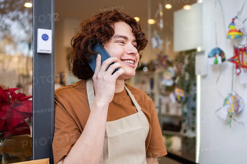 A cheerful shop owner wearing an apron stands at the organic products store entrance, smiling while engaging in a phone conversation The shop shop interior is visible behind them