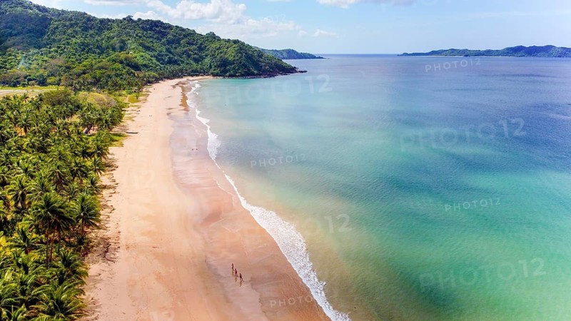 Aerial view of a serene beach with a lush green backdrop in the Philippines. With unrecognizable people walking along the shoreline, emphasizing the majestic scale of the natural scenery