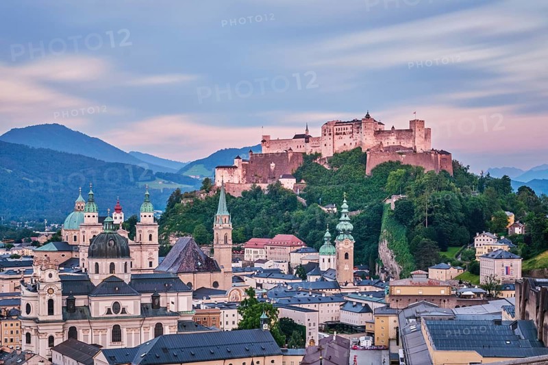 Long exposure aerial view of the illuminated Hohensalzburg Fortress and Salzburg Cathedral under a soft twilight sky at the end of the day