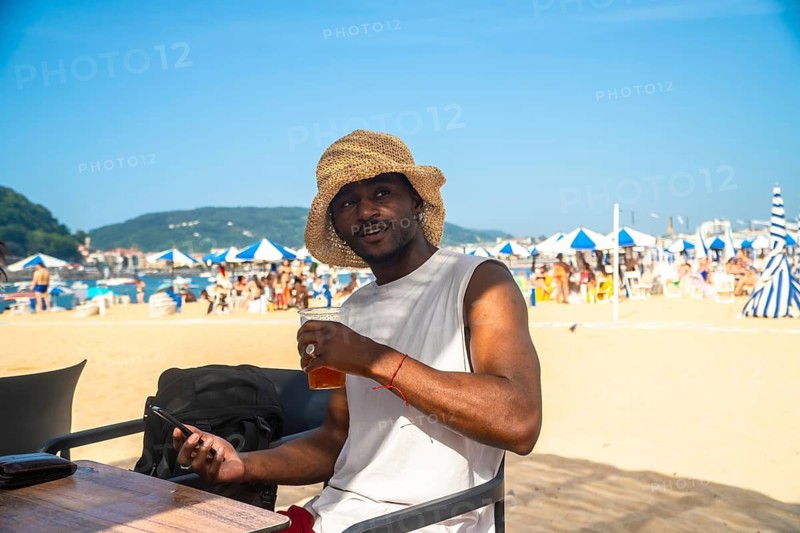 Young black man wearing a straw hat and white tank top enjoying a drink at a beach bar. Smiling and holding a smartphone