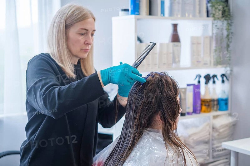 A hair stylist performs a keratin hair straightening treatment. The client sits in a salon chair wearing a cape while getting her hair brushed and prepared for the process