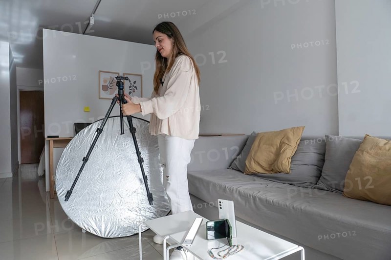Side view of woman adjusting camera tripod legs in living room, with silver reflector and phone stand with beaded strap visible in the background