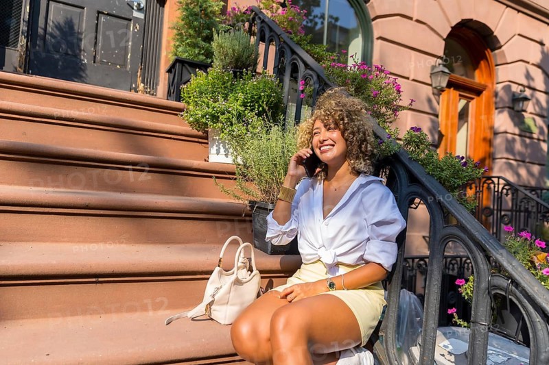Woman with curly hair sitting on steps of a new york city brownstone, smiling and talking on a mobile phone, embracing a relaxed urban lifestyle in the summer sun