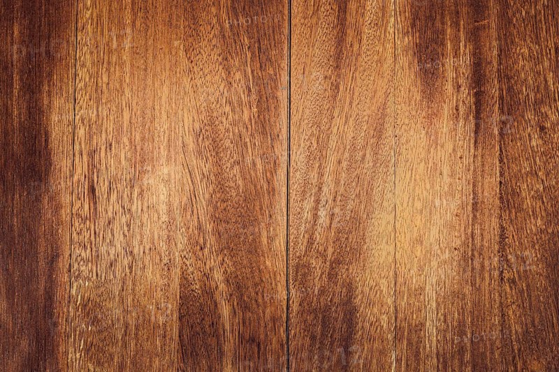 Close-up of a wooden surface with rich brown tones and visible grain patterns. The texture is smooth and polished, showcasing natural wood characteristics