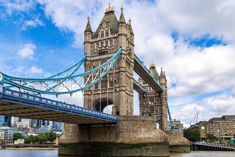 Looking up at Tower Bridge from the riverside path, the ornate stonework of the tower and its elevated blue walkway stand against scattered clouds, London, England, United Kingdom