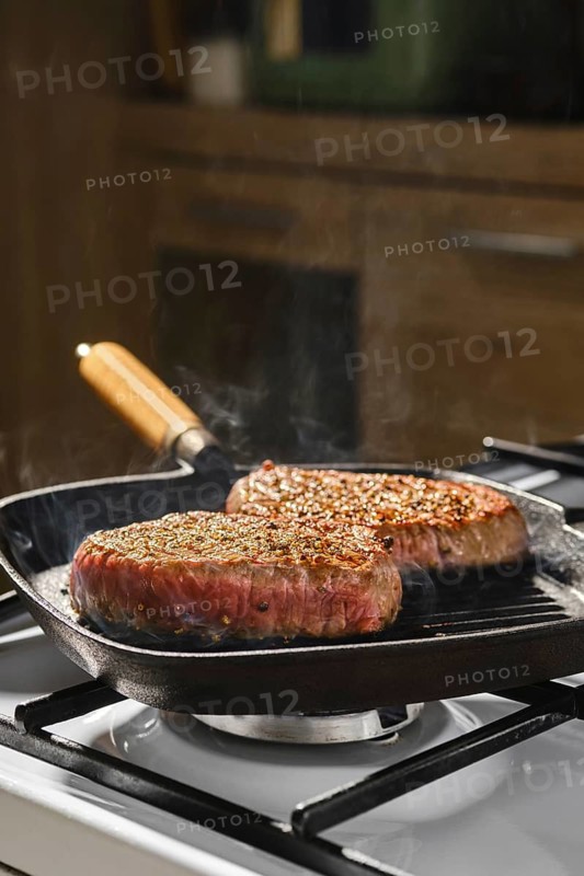 Two beef steaks are cooking on cast iron grill pan on gas stove in kitchen. Steam rises from meat as it cooks