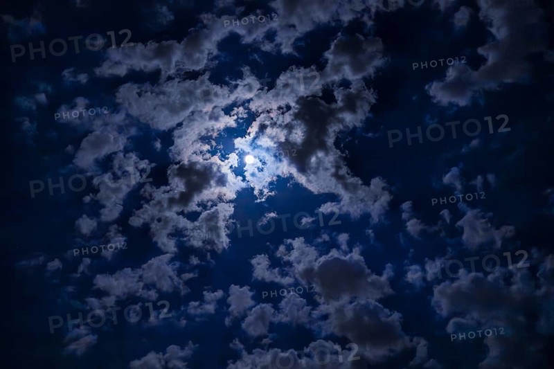 A night sky filled with dark clouds partially illuminated by a bright moon. The scene captures the beauty of nature at night
