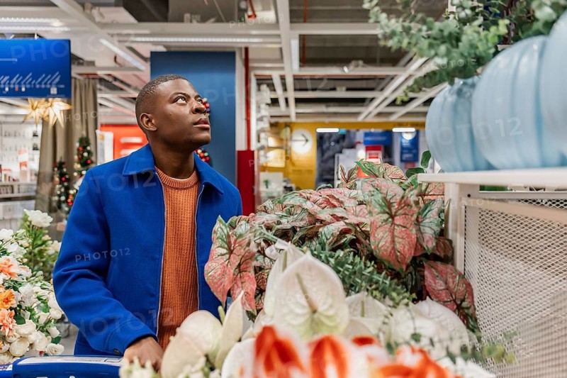 A young Black man with short hair wearing a blue coat and orange sweater looks up while shopping in a store filled with decorative plants and flowers
