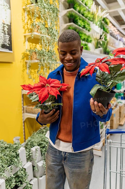 A person stands in a home goods store during the afternoon and arranges a red flower pot on a shelf. The store is filled with various plants and decorations