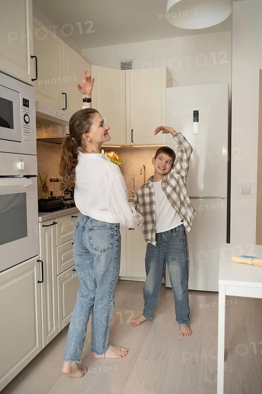 A joyful mother and son share a playful dance in a bright kitchen, creating a warm, heartfelt moment. The room is filled with natural light and familial happiness