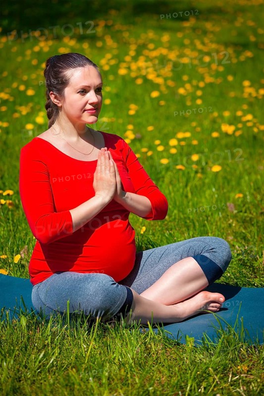 Pregnancy yoga exercise, pregnant woman doing asana Sukhasana easy yoga pose with namaste salutation outdoors on grass lawn with dandelions in summer