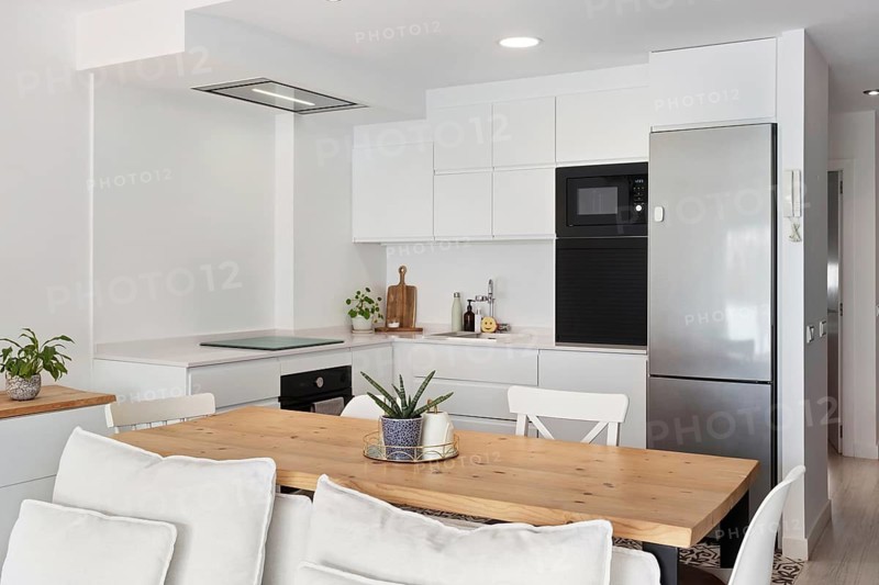 Bright and modern kitchen with sleek white cabinets and stainless steel appliances in the city center. A wooden dining table adds warmth, complemented by decorative plants and cozy seating
