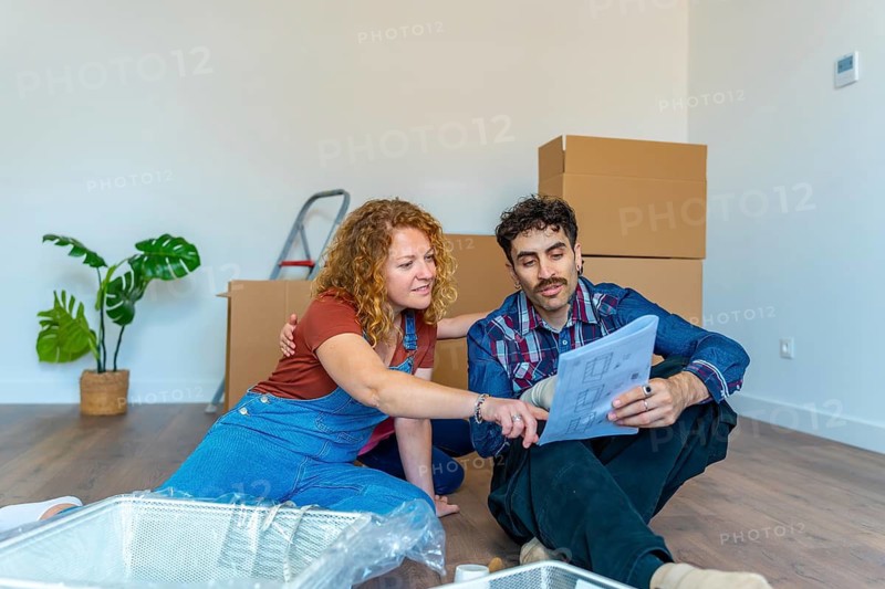 Young couple sitting on the floor, reviewing architectural plans in their new apartment surrounded by cardboard boxes, planning future home improvements
