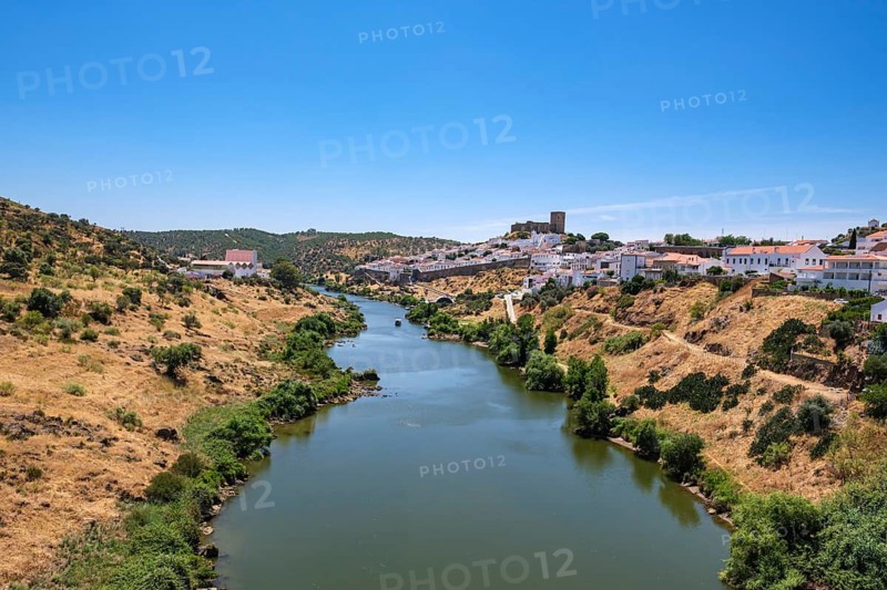 A calm river crosses a hilly landscape with a village on the shore under clear skies, Mértola, Beja, Portugal