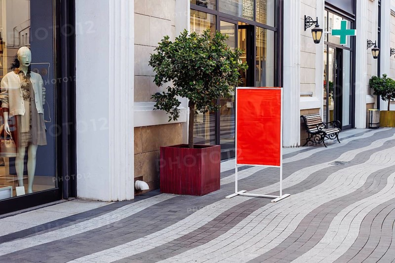 Mockup, retail storefront features bright red sign placed on clean street. Large windows showcase stylish clothing, while cheerful tree and bench encourage relaxation in this vibrant shopping area