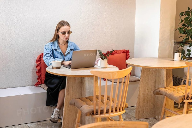 A young Caucasian woman with long blonde hair works on a laptop at a cafe table. She wears sunglasses and has a cup of coffee beside her