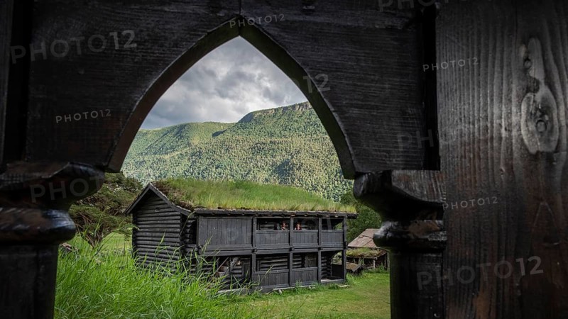 View through a wooden archway, traditional wooden house with grass sod roof in front of a cloudy mountain landscape, backdrop Kristin Lavranstochter film, Jørundgard Middelaldersenter, Olavsweg or Olavsleden, Sel municipality, Gudbrandsdalen, Norway