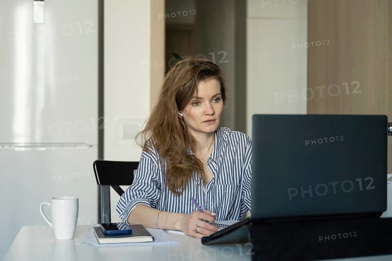 Woman in striped shirt works at home, focused on laptop. She writes in a notebook with a cup on the table, symbolizing work-from-home productivity