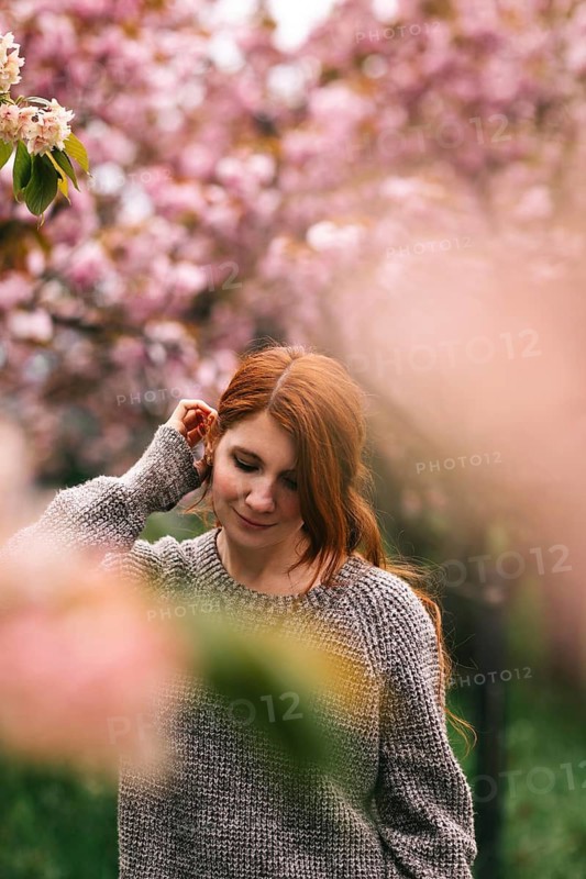 A young woman with red hair enjoys a serene moment in a lush garden surrounded by blooming tulips and cherry blossoms. Her peaceful expression complements the vibrant spring setting