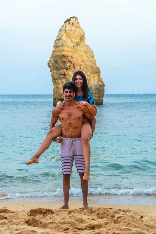 Tourists are having fun on a beautiful beach in the algarve region of portugal, with the man giving his partner a piggyback ride in front of a distinctive rock formation