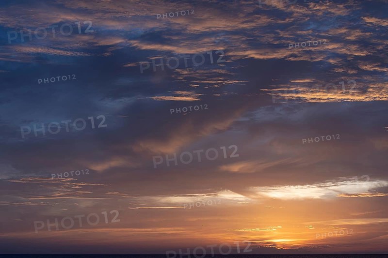 A serene sunset over the sea with a vibrant display of clouds catching the warm hues of the setting sun. The sky shifts from deep blue to vibrant oranges and yellows