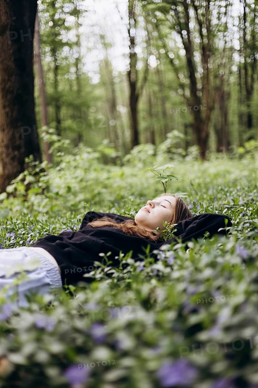 A serene teenage girl lies on a bed of lush periwinkle flowers, her eyes closed, in a tranquil forest setting, capturing the essence of nature's peace and beauty