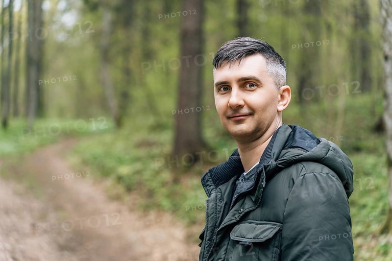 A man wearing a dark jacket walks along a forest trail surrounded by tall green trees and dense foliage. The curved path leads deeper into the woods, creating a peaceful outdoor moment that highlights nature, solitude and the calm atmosphere of a quiet forest