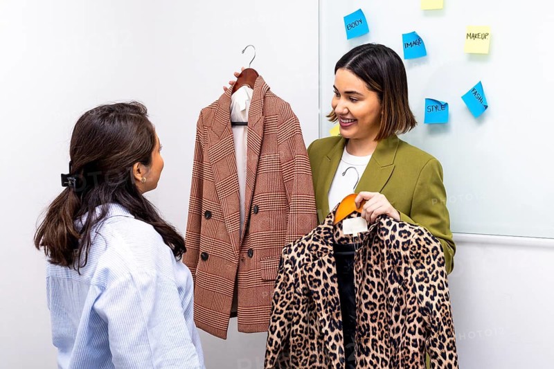 A fashion consultant assists a woman in selecting the perfect outfit by comparing a plaid blazer and a leopard print jacket. Sticky notes on the wall highlight style tips