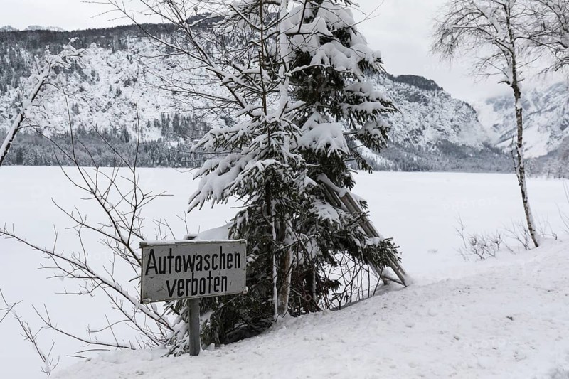 German language sign not to wash your car in the lake. Snow covered Lake Almsee which is partly frozen and snow covered. Snow on the trees and mountains with gray overcast sky. Nearly a black and white scene