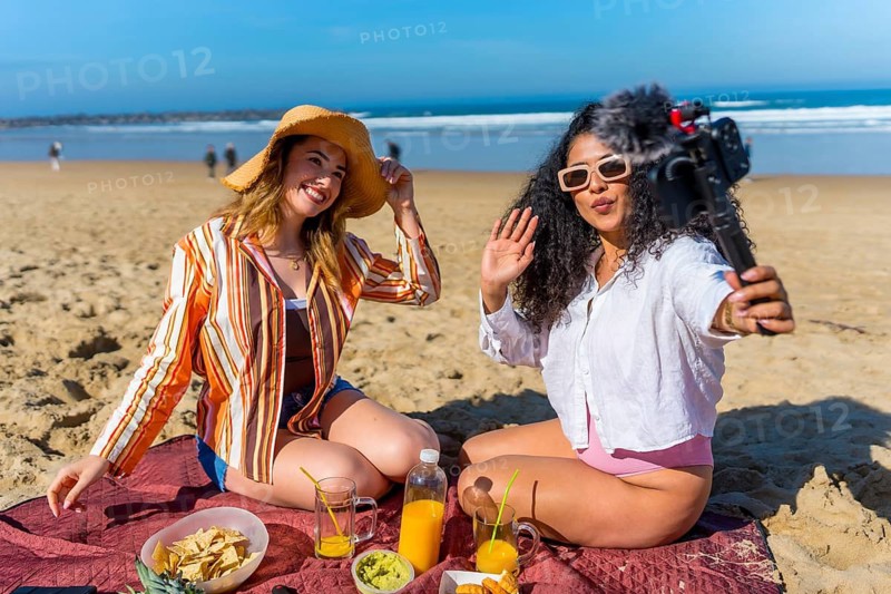 Two young women enjoying a beach picnic, vlogging their fun summer vacation experience and actively creating content for social media, capturing moments with a camera