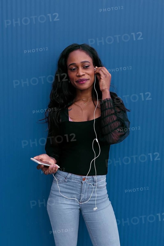 A young woman stands against a vibrant blue background, smiling as she listens to music through earphones connected to her smartphone, conveying a mood of relaxation and enjoyment