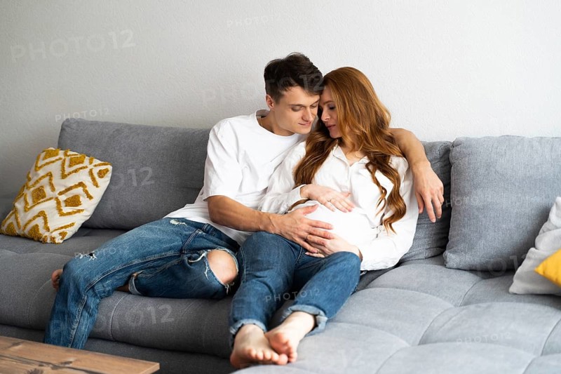 A young couple sits closely on a comfortable couch, with the man gently touching the woman's pregnant belly. They appear relaxed and happy, embracing the journey of parenthood