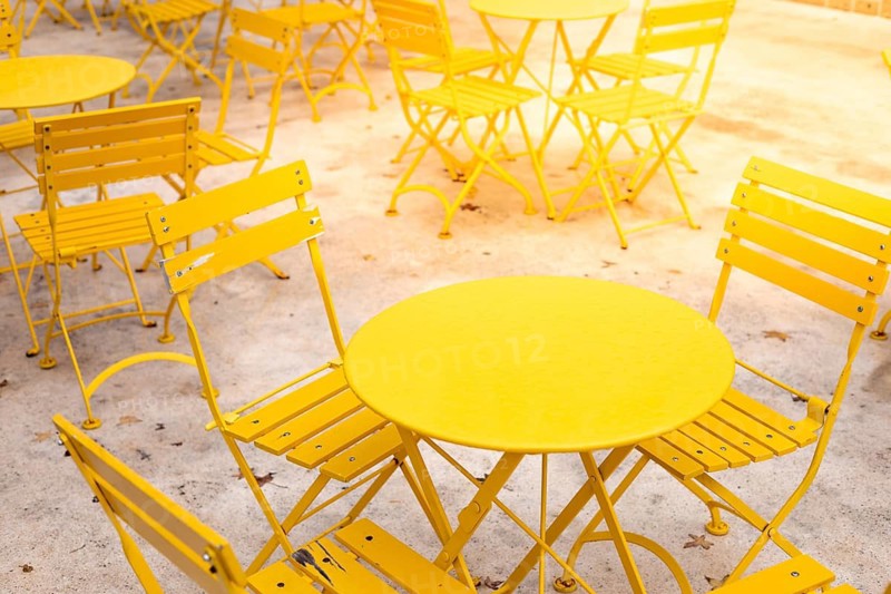 Rows of bright yellow metal tables and chairs are arranged in an outdoor area. The sunny environment enhances the cheerful vibe. Icecream, coffe and dessert summer cafe style