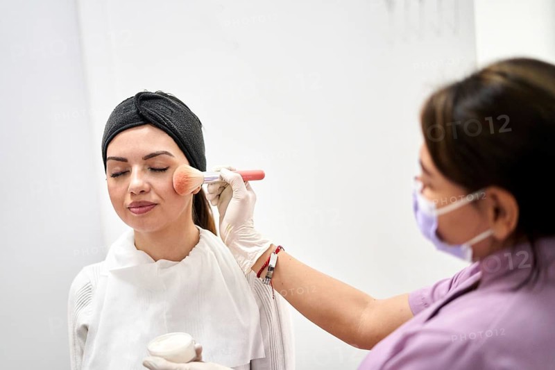 A beauty specialist applies makeup using a brush on a client who is enjoying a facial treatment. The setting is a professional aesthetic medicine clinic focused on relaxation and skincare