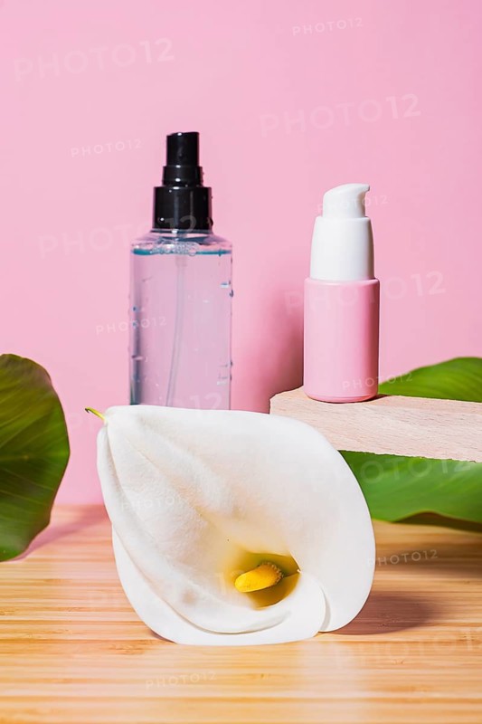 Two modern cosmetic containers sit on a wooden platform with a pristine white calla lily, over a pink background highlighting fresh skincare presentation