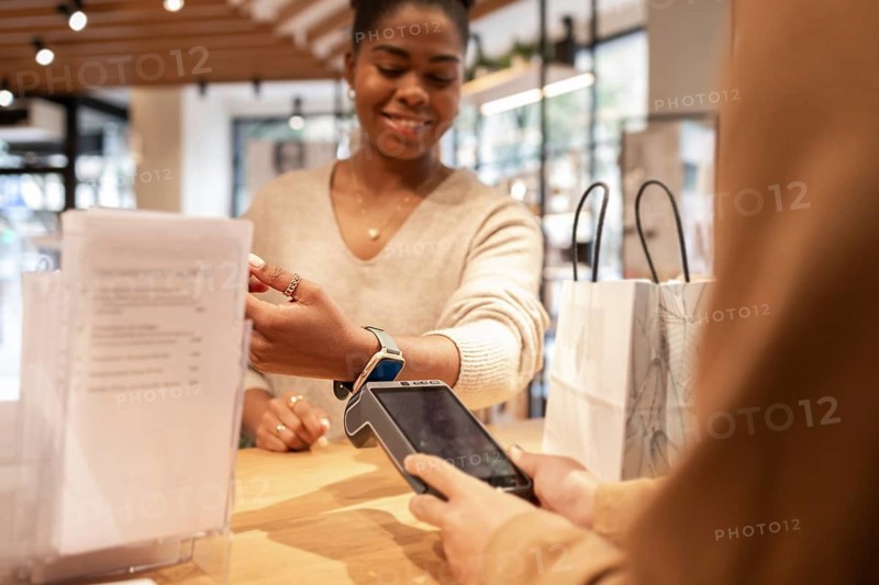 A customer smiles while making a contactless payment with a smartwatch in a trendy cosmetics store. Shopping bags and product brochures are visible on the counter